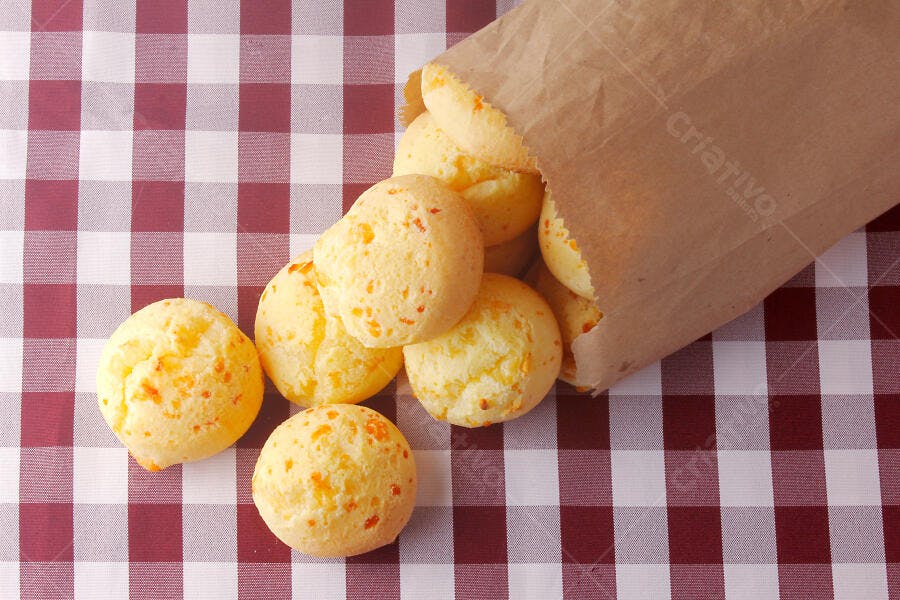 Pão de queijo quente saindo de saco de papel sobre toalha xadrez vermelha e branca