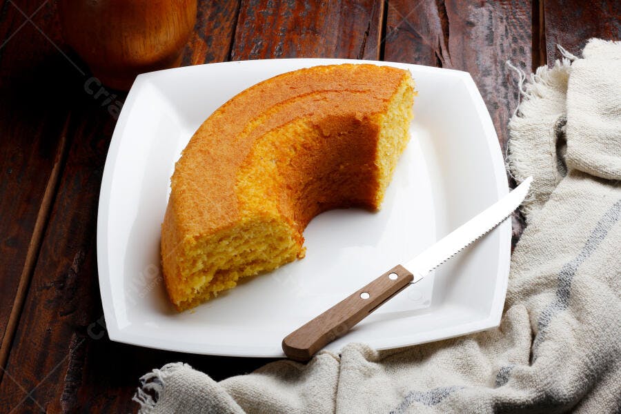 Fatia de bolo de fubá amarelo em prato branco com faca sobre mesa de madeira rústica