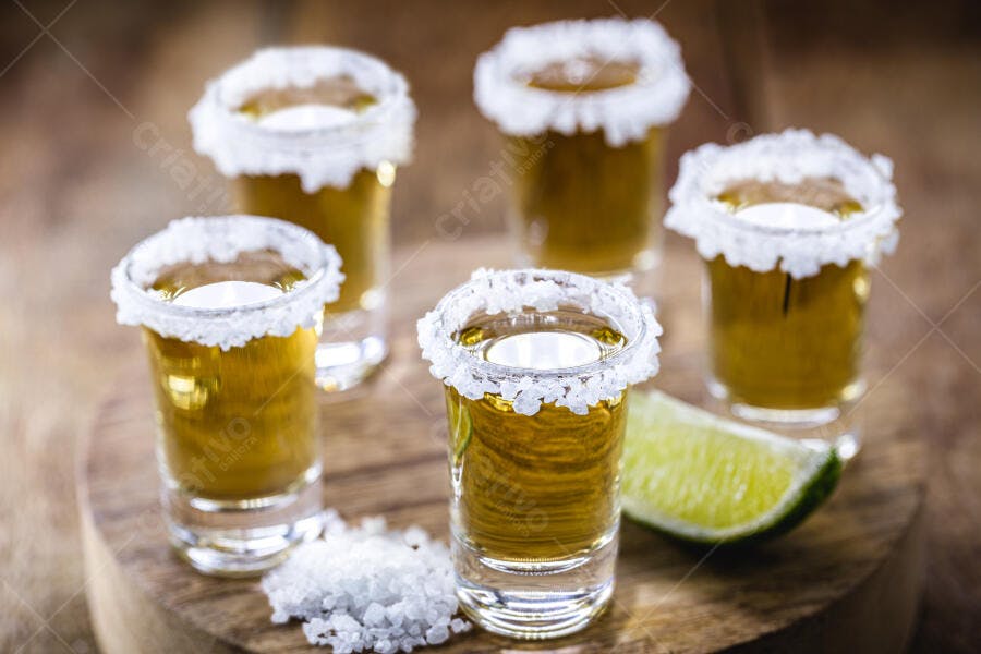 Shots de tequila dourada com borda de sal e limão fresco em tábua de madeira