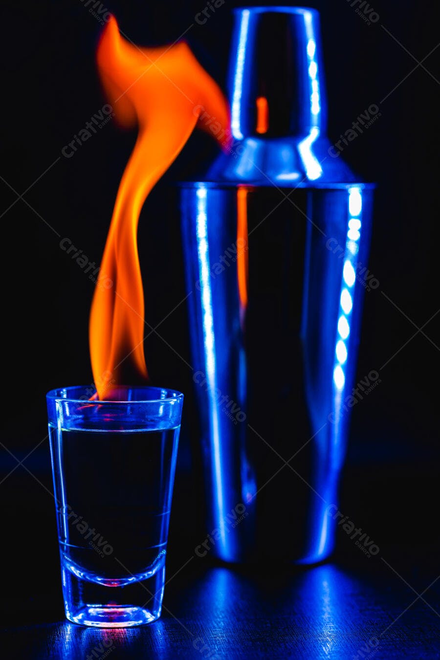 Copo de shot com chama laranja e coqueteleira azul neon em ambiente escuro