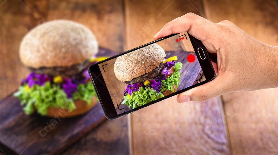 Mão filmando hambúrguer gourmet colorido com telefone celular em mesa de madeira