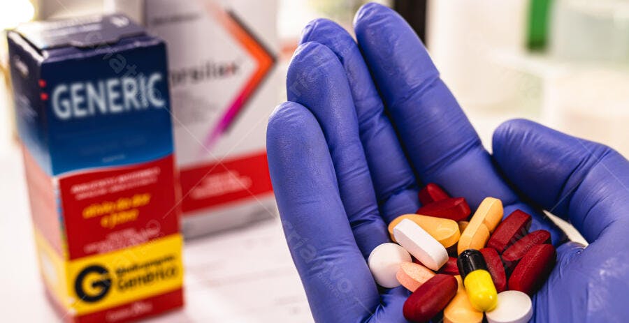 Mão com luva azul segurando pílulas coloridas e medicamentos genéricos