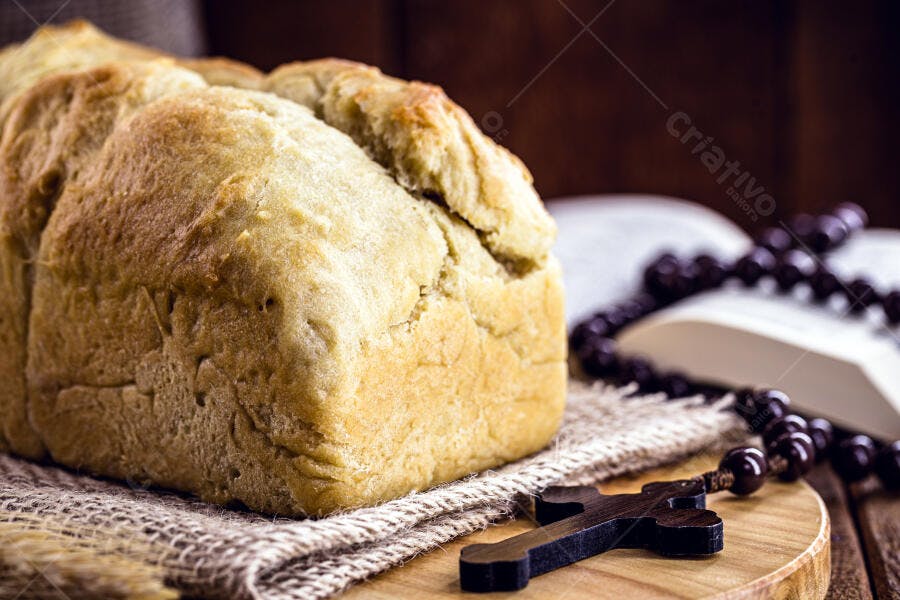 Pão caseiro dourado com rosário e bíblia aberta em mesa de madeira