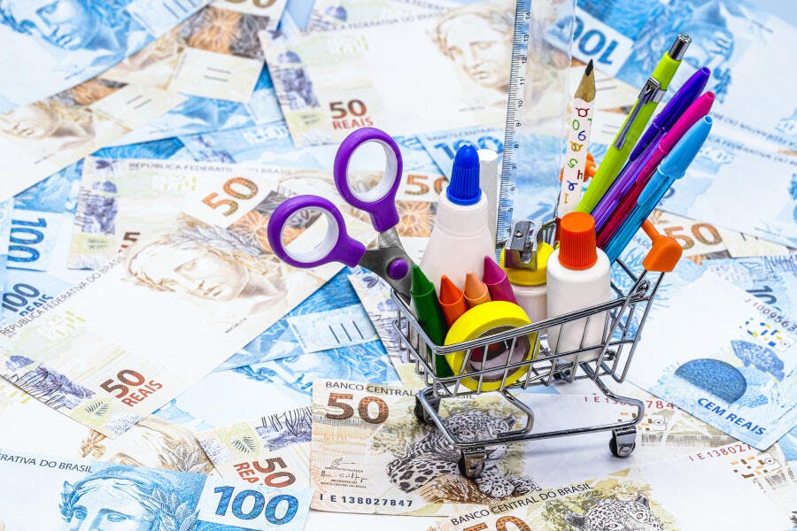 Materiais escolares coloridos em carrinho de supermercado pequeno sobre notas de Real