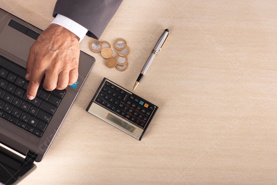 Homem de negócios digitando no laptop com moedas e calculadora em mesa bege