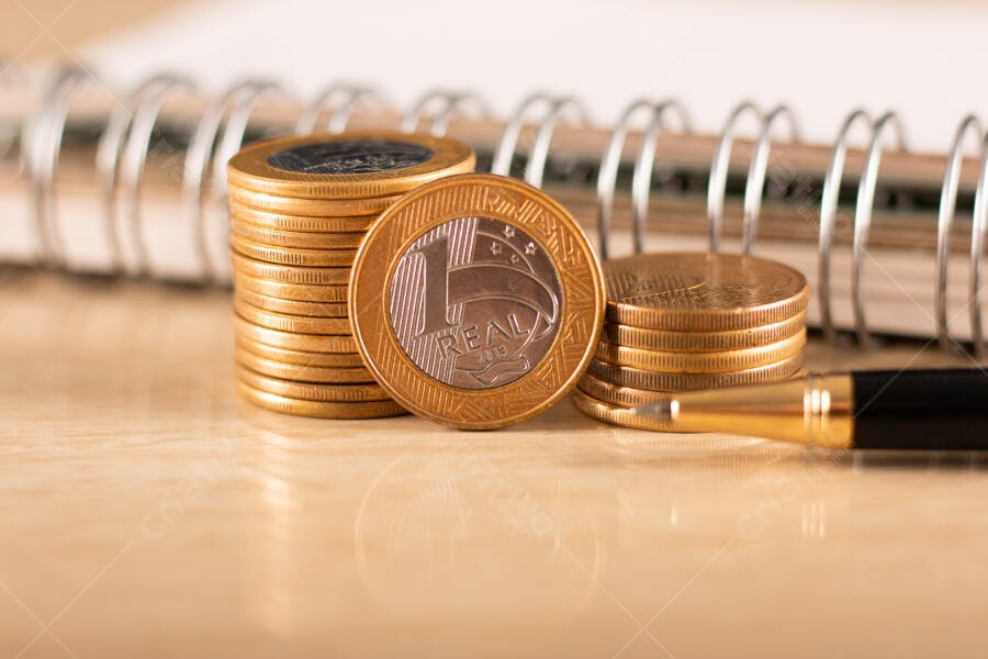 Moedas de Real douradas empilhadas sobre mesa com notebook e caneta