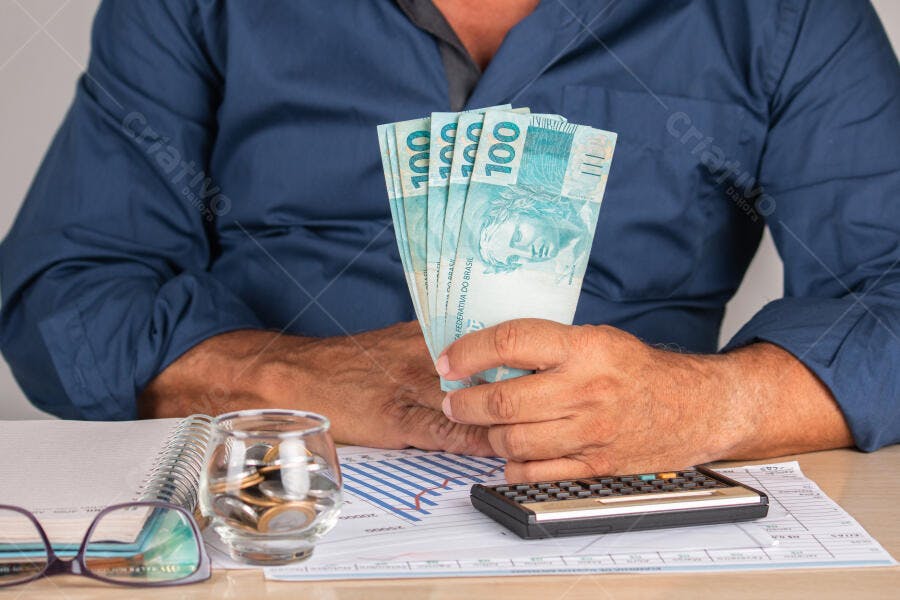 Homem segurando notas de cem reais em mesa de escritório com calculadora