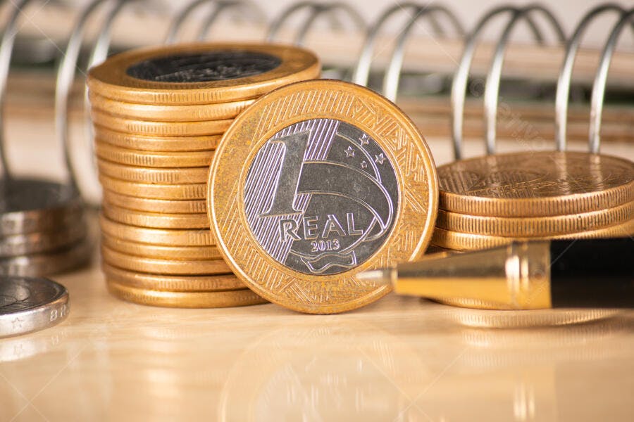 Moedas de Real empilhadas em mesa com caderno finanças e economia