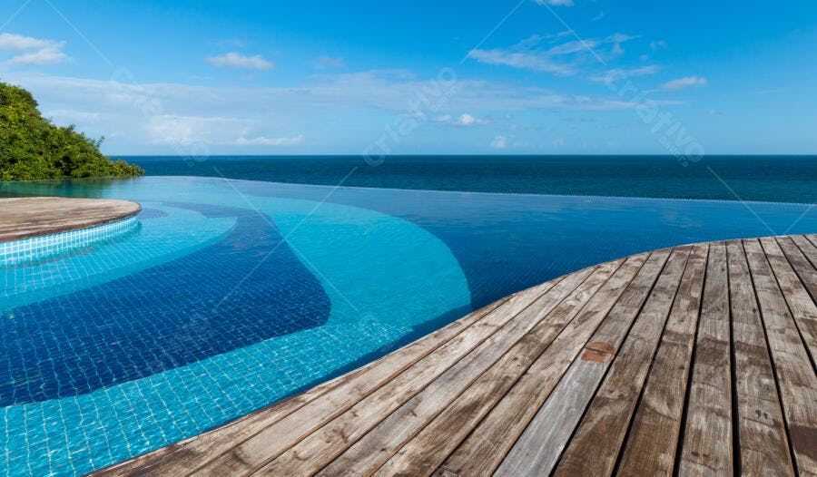 Piscina de borda infinita com vista para oceano azul e deck de madeira em dia ensolarado