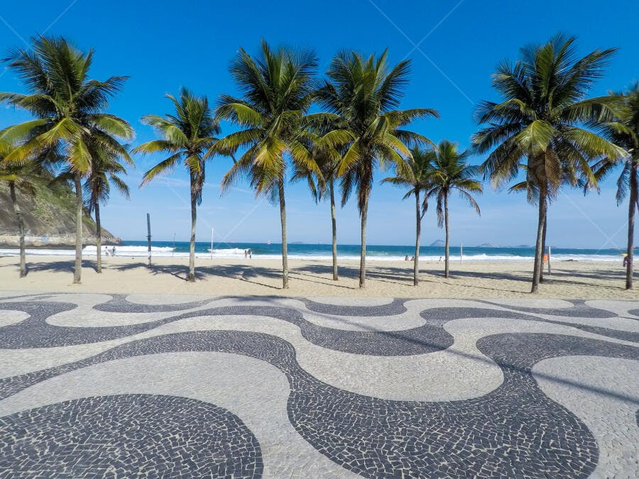 Praia de Copacabana com calçadão e coqueiros sob céu azul em dia ensolarado no Rio