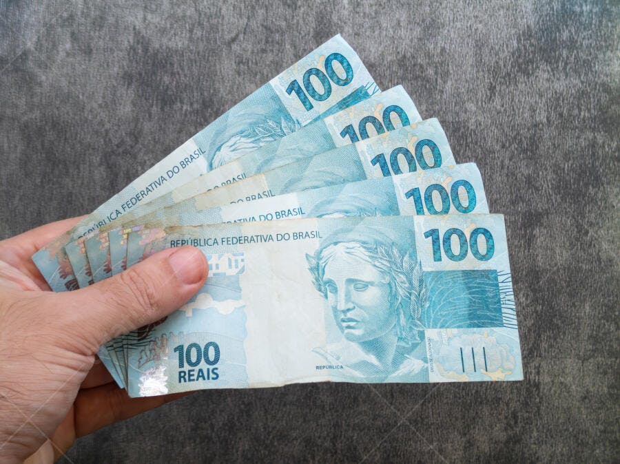 Mão segurando cédulas azuis de 100 Reais moeda brasileira em fundo cinza