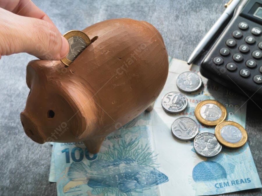 Mão depositando moeda de Real em cofrinho de barro com notas e calculadora