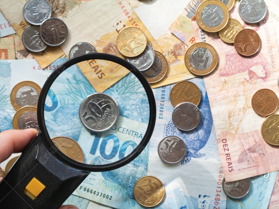 Moedas e notas brasileiras sendo examinadas por lupa em tons de azul rosa e dourado