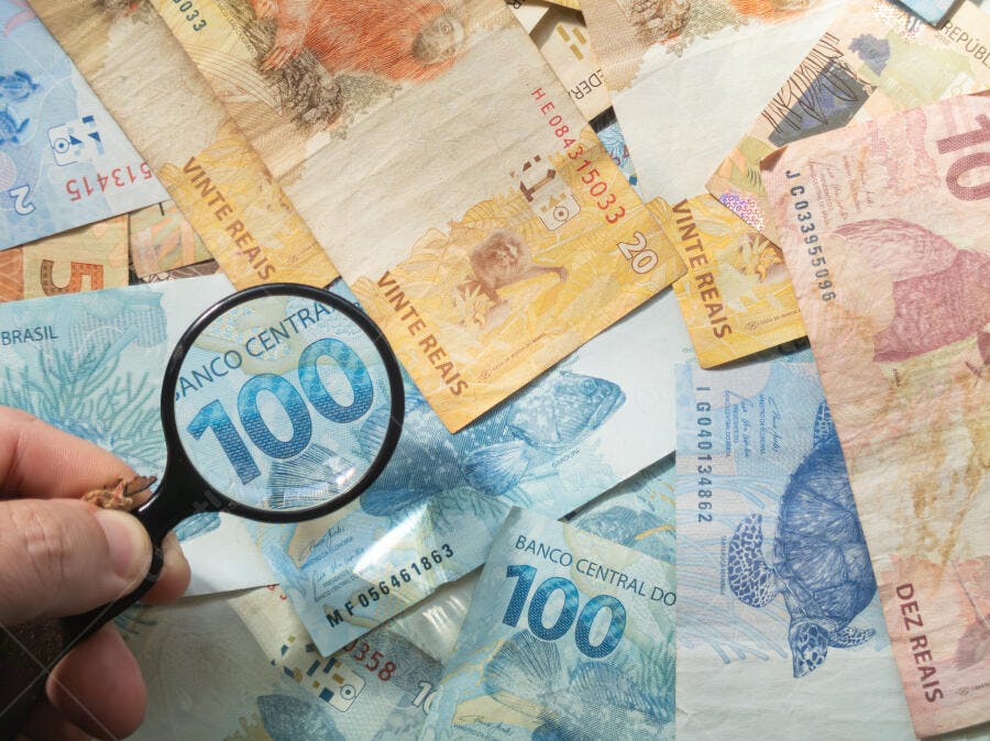 Mão com lupa examinando notas de reais brasileiras coloridas de 100 e 20