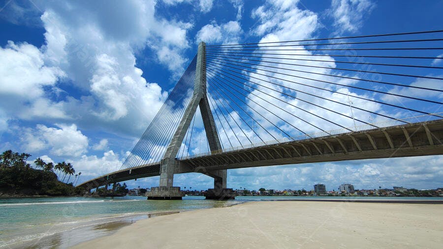 Ponte estaiada cinza sobre praia tropical com céu azul e nuvens brancas