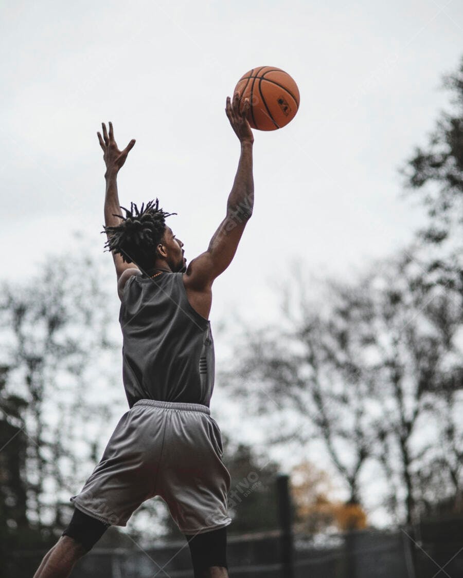 Homem negro atlético jogando basquete em quadra externa pulando para arremessar a bola
