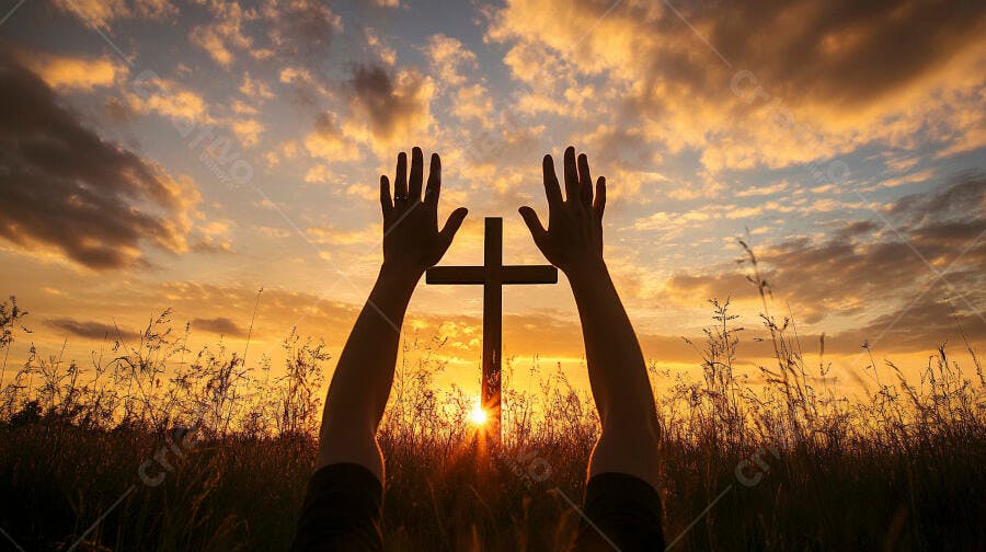 Mãos levantadas em oração e cruz ao pôr do sol em campo de grama