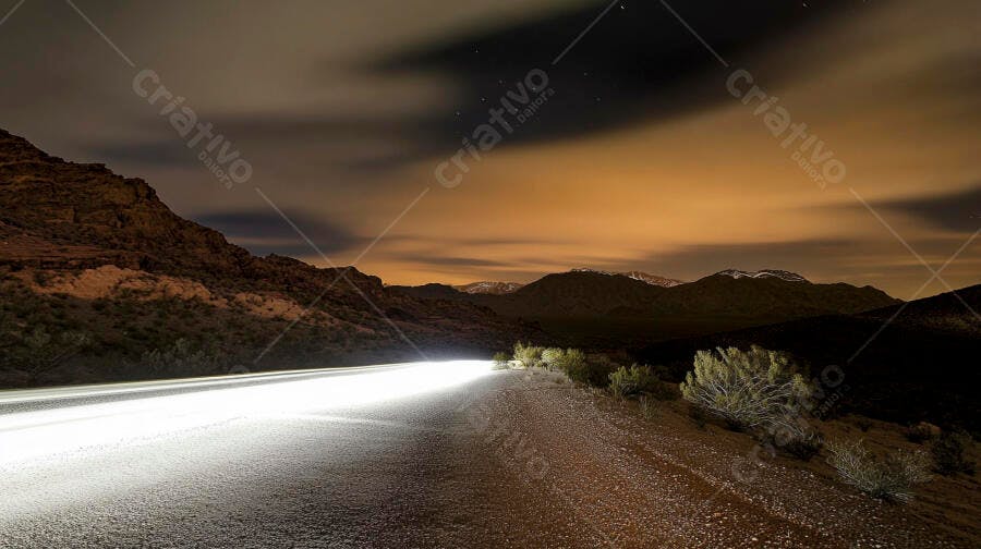 Estrada do deserto noturna com rastro de luz de carro sob céu laranja com montanhas