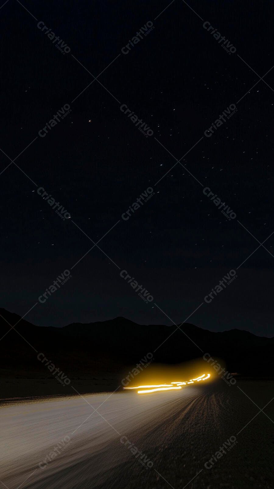 Rastros de luz amarelo em estrada noturna sob céu estrelado e montanhas distantes
