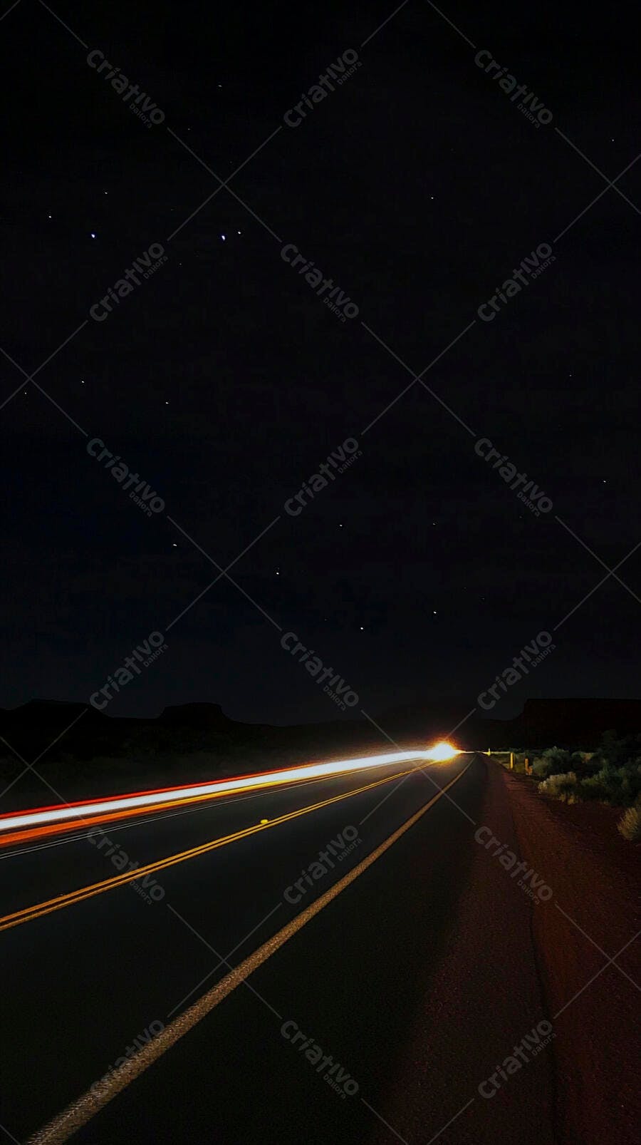 Rastros de luz de veículos em estrada noturna sob céu estrelado e paisagem escura