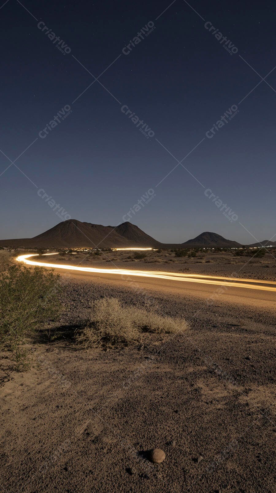 Estrada noturna deserta com rastros de luz de carros e montanhas sob céu estrelado