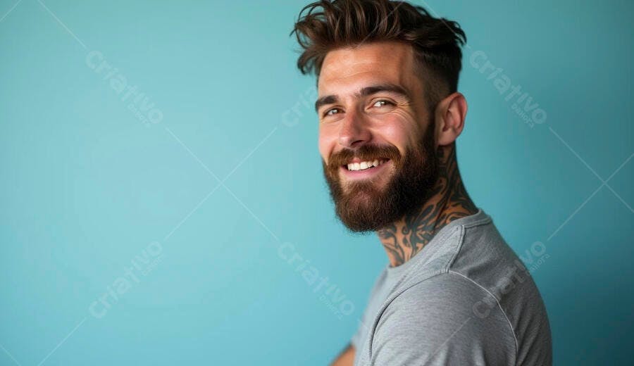 Homem caucasiano barbudo sorrindo com tatuagens e camiseta cinza em fundo azul