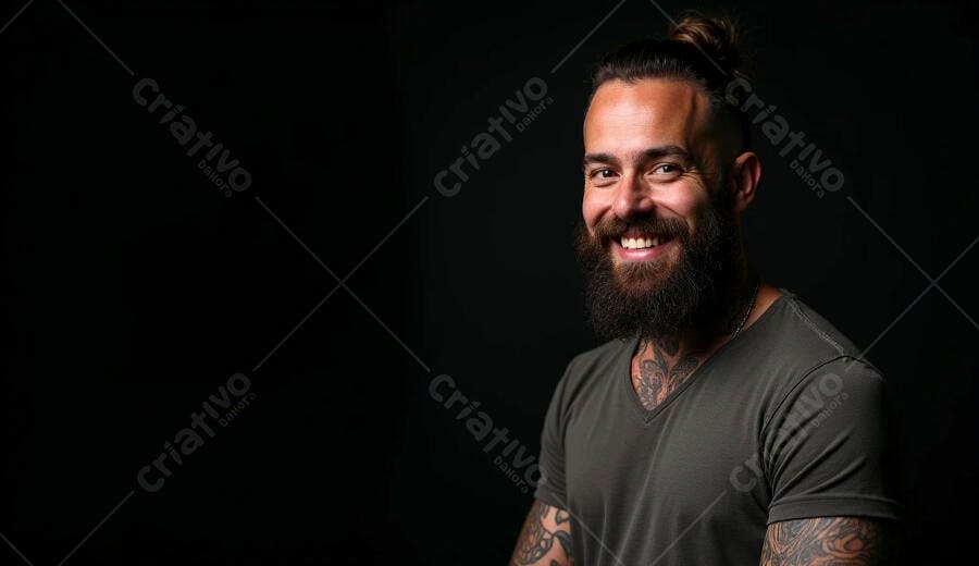 Homem barbudo sorrindo feliz com tatuagens cabelo coque e camiseta verde em estúdio escuro