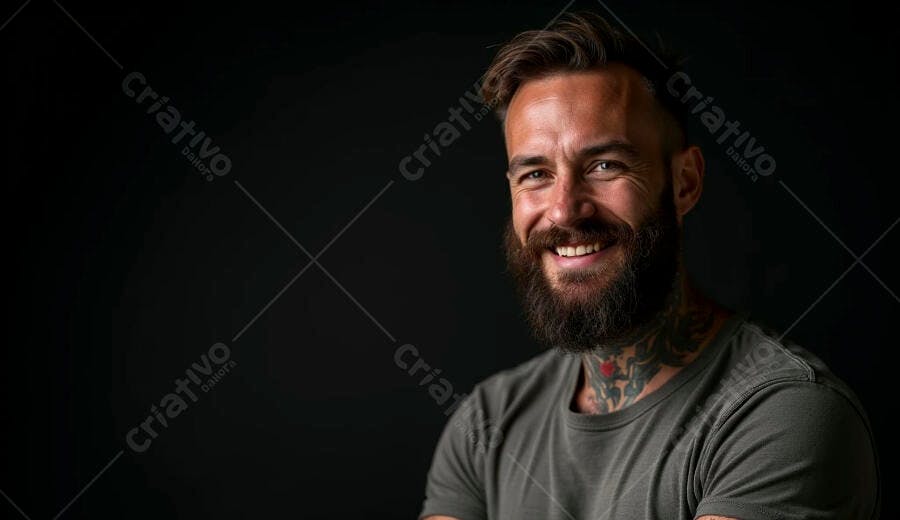 Homem barbudo sorrindo com tatuagens e camiseta cinza em fundo escuro