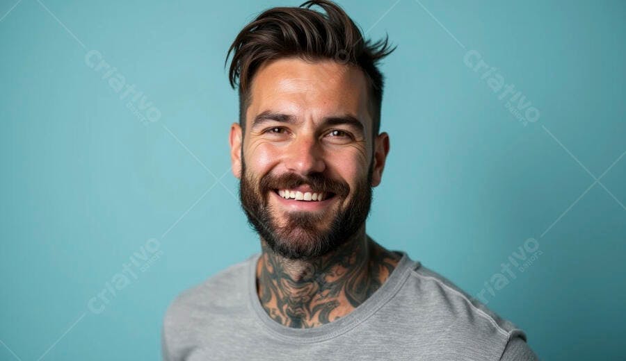 Homem barbudo tatuado sorrindo com camisa cinza em fundo azul claro